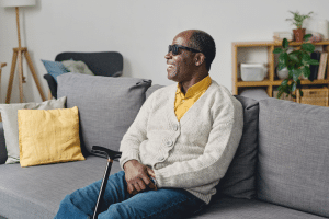 A imagem mostra um homem idoso cego, sentando em um sofá, sorrindo, enquanto sua bengala está apoiada em uma de suas pernas. Ele utiliza óculos escuros, calça jeans e um suéter branco por cima de uma camisa amarela e está em sua sala e ilustra o texto 