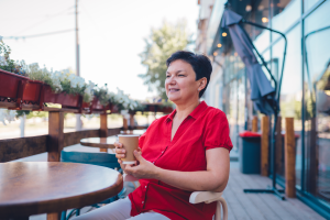 A imagem mostra uma mulher madura em um café, sentada em uma cadeira, enquanto sorri e segura um copo de café e ilustra o texto INSS e aposentadoria: pedidos, como reverter negativa e receber da Koetz Advocacia.