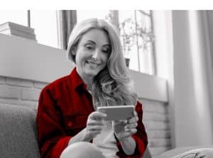 A imagem mostra uma mulher madura alegre, enquanto navega na internet em seu celular. A imagem ilustra o texto 