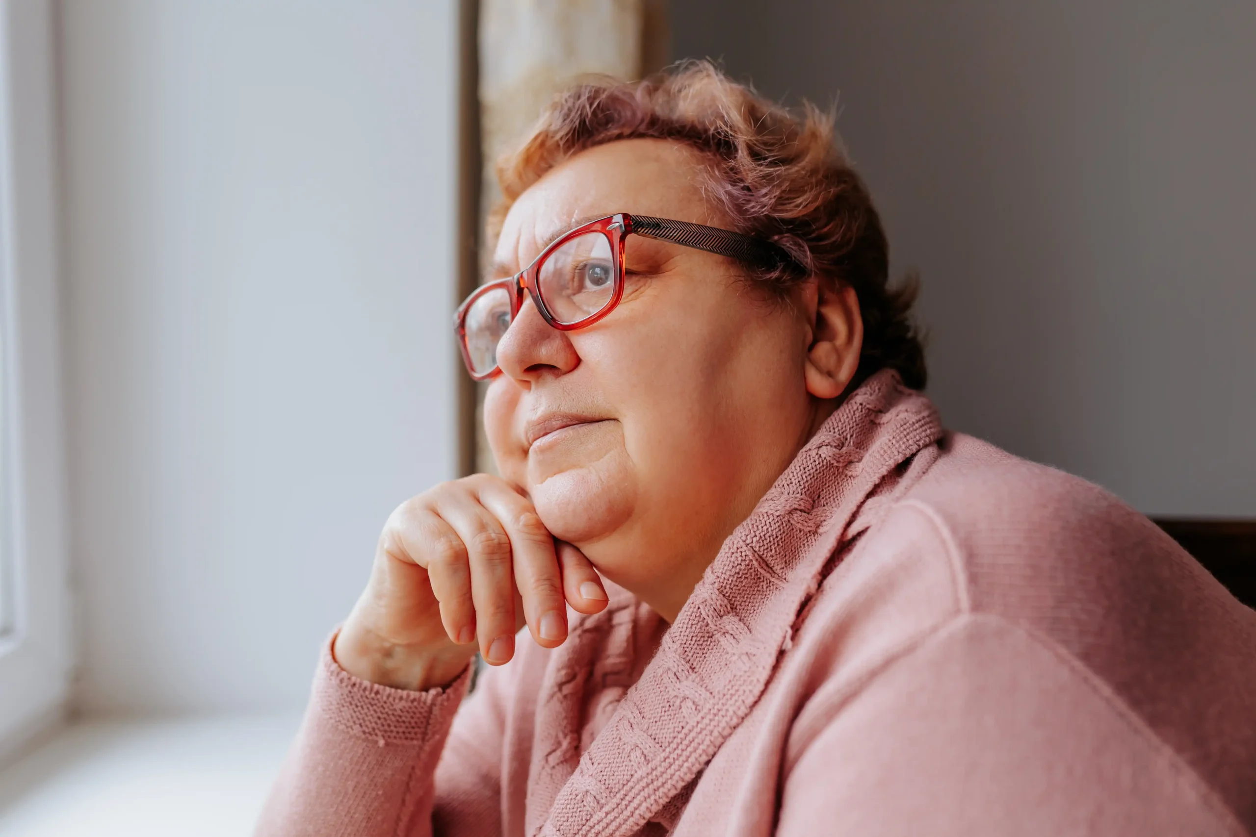 A imagem mostra uma mulher sênior pensativa, com uma das mãos no queixo. Ela possui cabelos curtos castanhos, usa óculos de grau com aro vermelho e um suéter rosa.