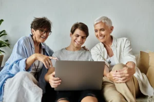 A imagem mostra uma família LGBTQ+ alegre, se unindo em casa em um momento aconchegante. O filho jovem adulto mostra algo em seu latptop para suas mães. Ilustra o texto sobre aposentadoria por pontos.