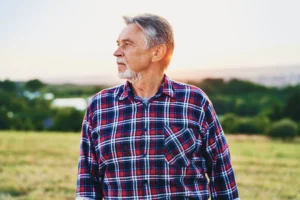 A imagem mostra um homem sênior, ao ar livre, olhando para o horizonte. Ele possui cabelos e barba grisalhos e usa uma camisa xadrez.