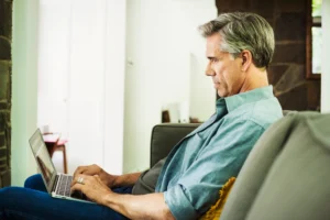 A imagem mostra um homem de meia-idade, sentado em um sofá, dentro de casa, utilizando o laptop.