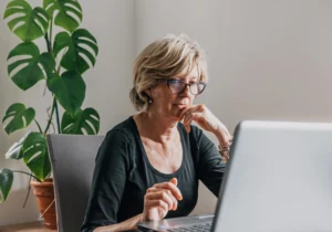A imagem mostra uma mulher de meia-idade, sentada à mesa, utilizando um laptop.