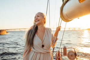 Mulher madura em segurando cordas de um barco em alto mar, sorrindo e com o pôr do sol ao fundo - a imagem ilustra o texto sobre Aposentadoria Internacional da Koetz Advocacia.