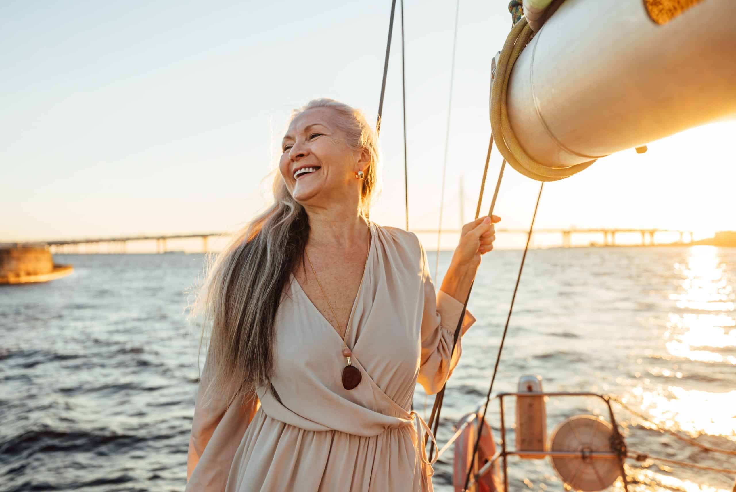 Mulher madura em segurando cordas de um barco em alto mar, sorrindo e com o pôr do sol ao fundo - a imagem ilustra o texto sobre Aposentadoria Internacional da Koetz Advocacia.