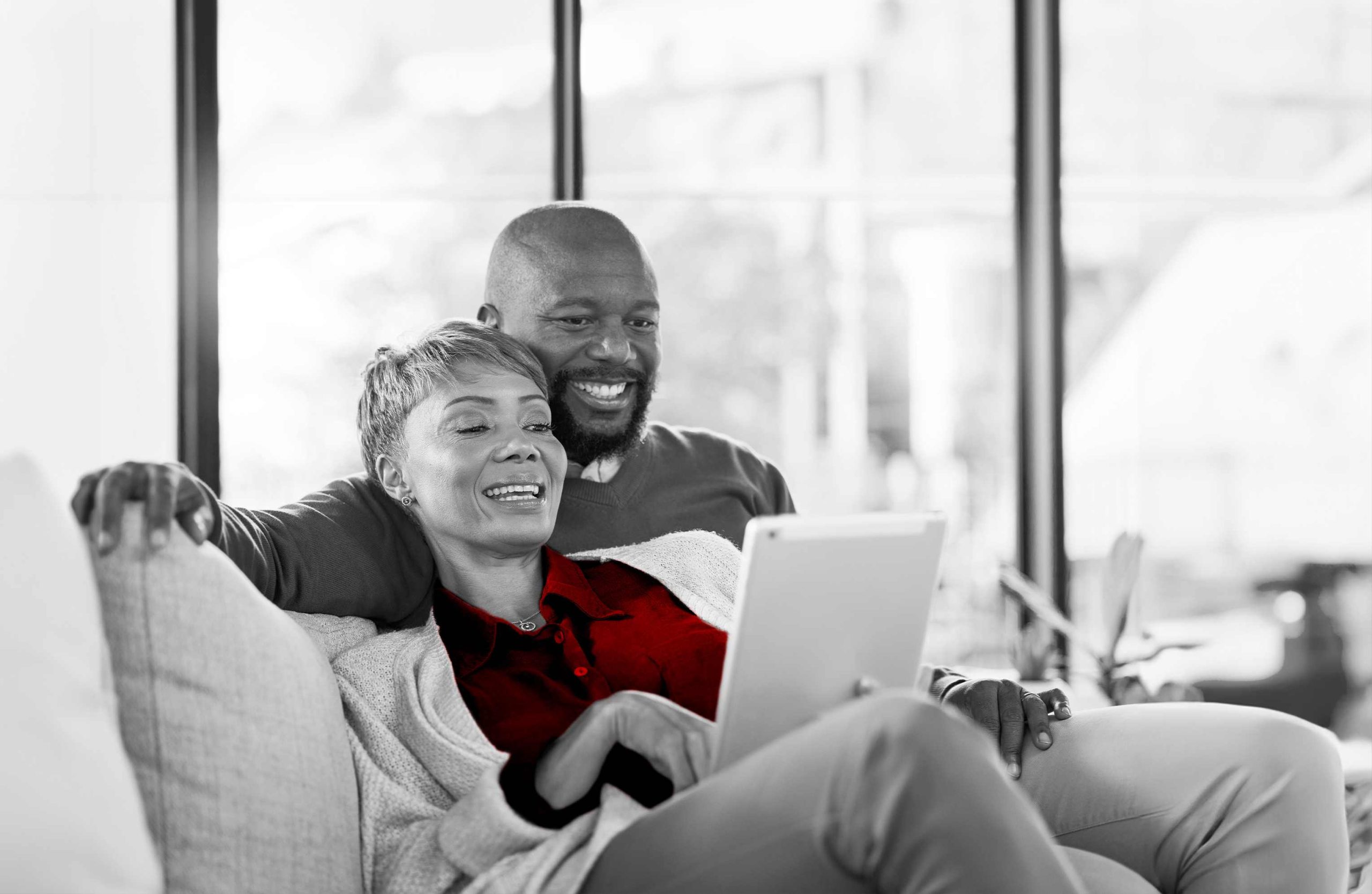 A imagem mostra um casal sorrindo no sofá, abraçados, olhando para o computador.