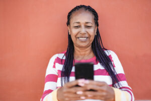 A imagem mostra uma mulher idosa, negra e com cabelos longos, encostada em uma parede vermelha, enquanto segura com as duas mãos um celular smartphone. Ela sorri, enquanto olha para a tela.