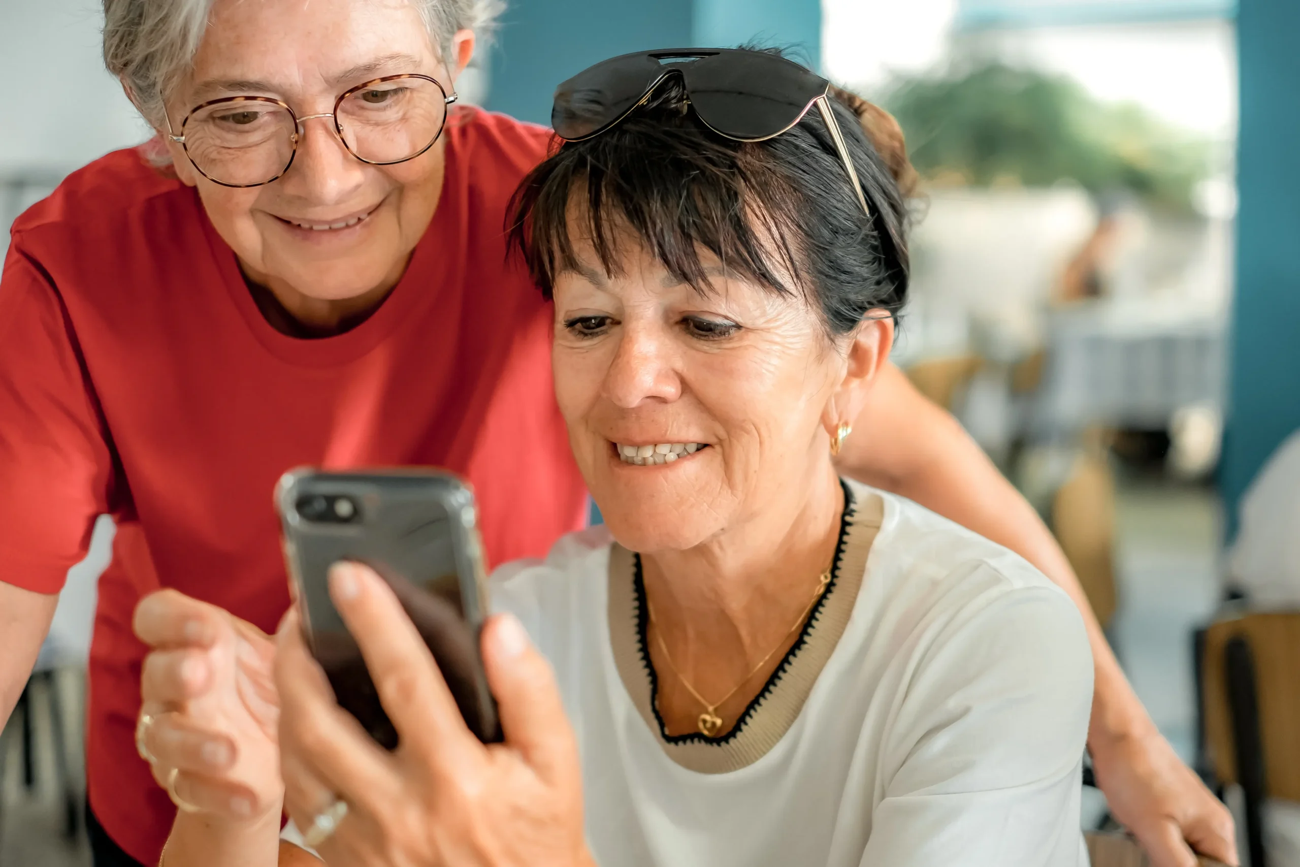 A imagem mostra duas mulheres idosas sorrindo, olhando para o celular. Uma possui cabelos curtos brancos, está usando uma roupa vermelha e a outra mulher, ao seu lado, possui cabelos curtos castanhos e segura o celular com uma das mãos.