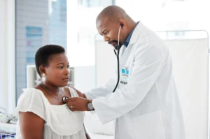 A imagem mostra uma médico, de jaleco, em abiente hospitalar, atendendo uma paciente.