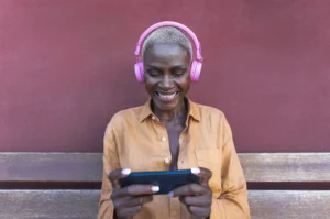 A imagem mostra uma mulher negra, madura, de cabelos curtos, usando smartphone móvel enquanto ouve música com fones de ouvido sem fio.