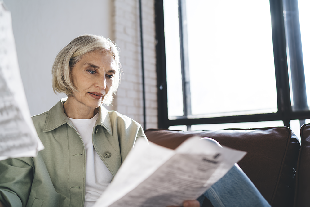 Mulher branca, loira, de aproximadamente 50 anos, sentada no sofá com documentos na mão. Ela olha para os documentos com uma expressão reflexiva e ilustra o texto Quanto pagar de INSS como autônomo