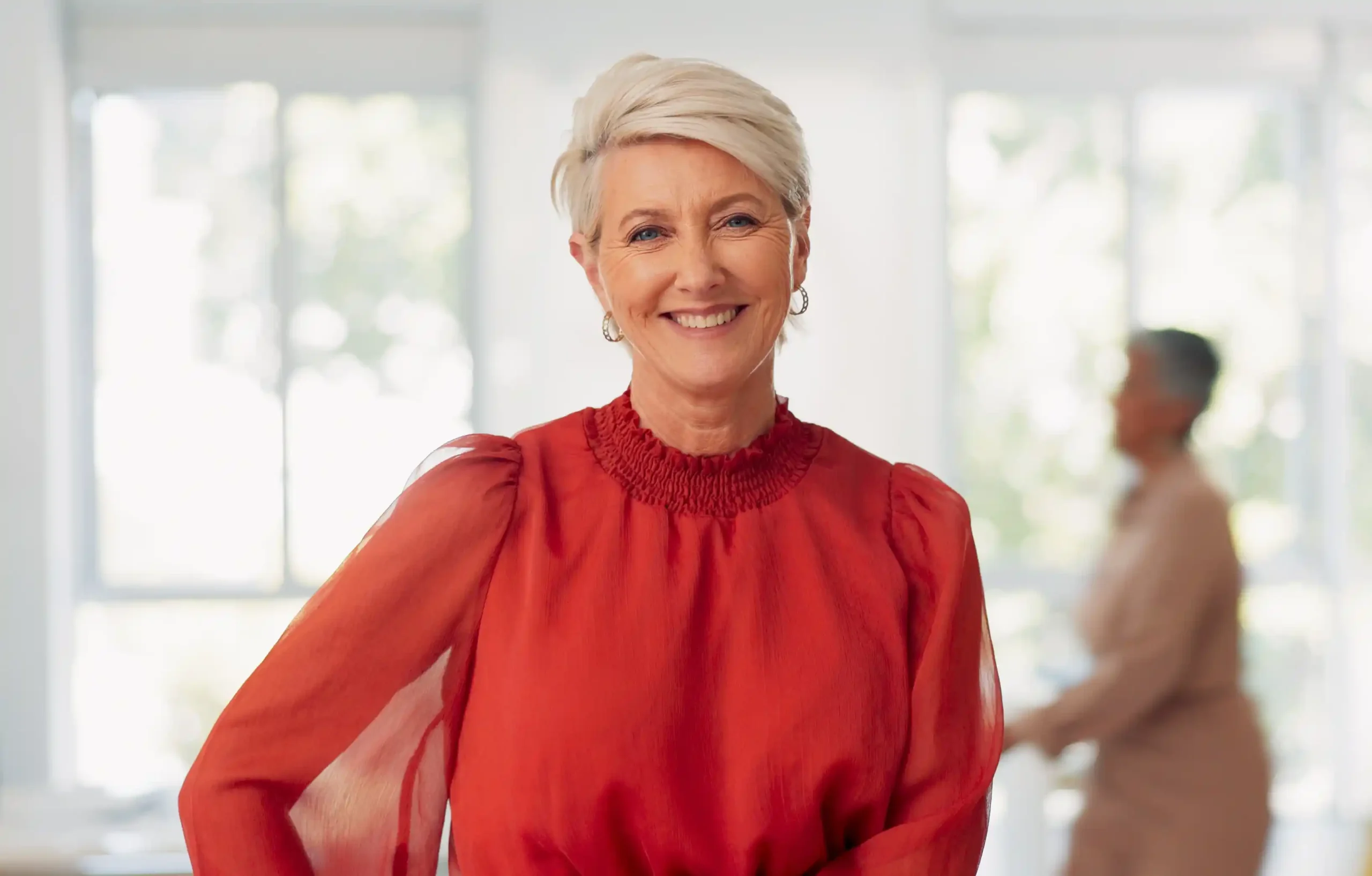 A imagem mostra uma mulher madura, de cabelos brancos e curtos, sorridente. Ela está olhando para a câmera, usando uma blusa vermelha e está em um ambiente de trabalho.