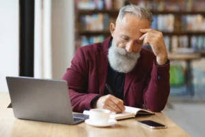 A imagem mostra um empresário sênior pensativo verificando sua agenda, trabalhando em um café. Ele possui cabelos e barbas brancas e está usando um terno vinho.