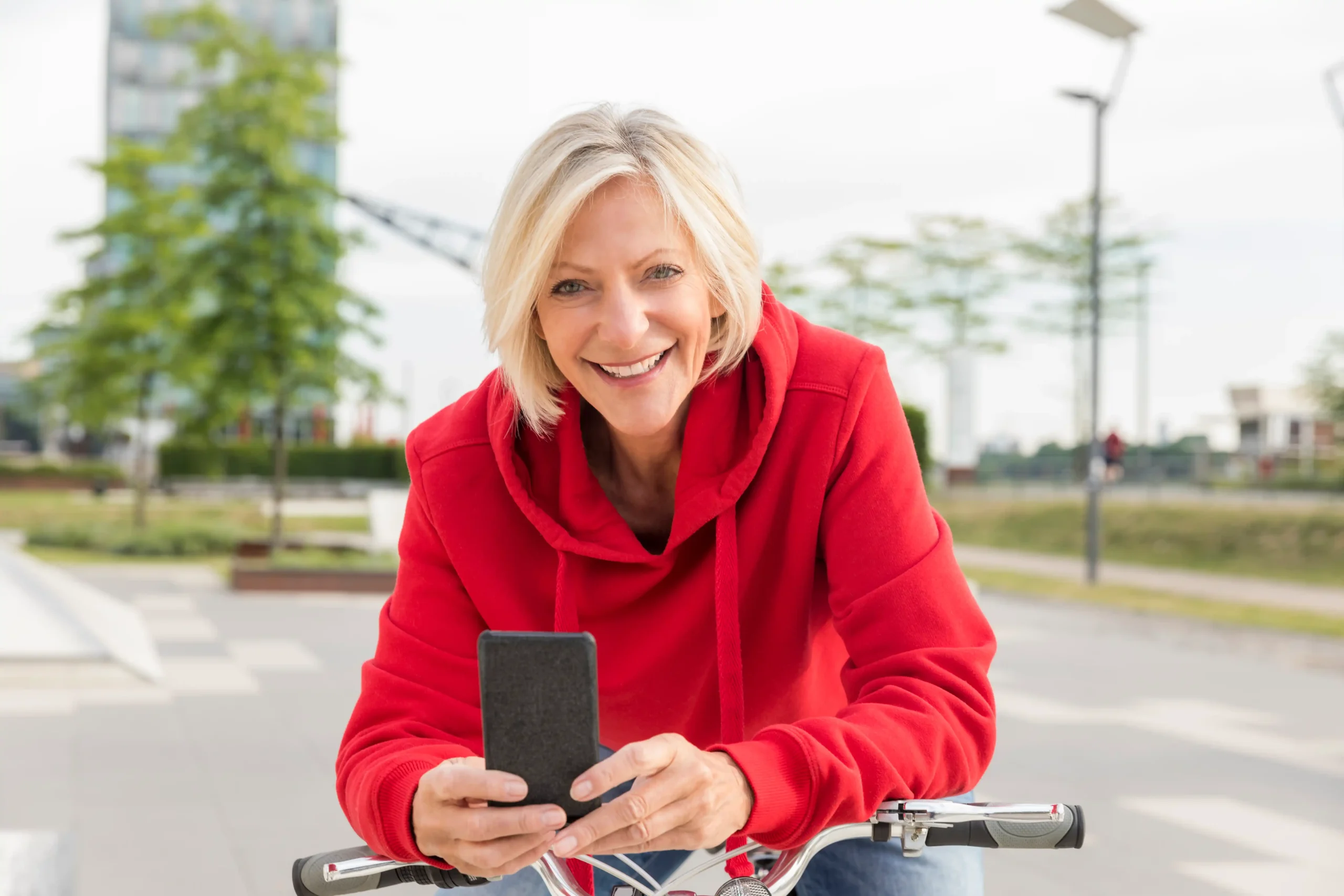 A imagem mostra uma mulher madura, andando de bicicleta pela cidade, enquanto segura um celular nas nãos. Ela tem cabelos médios loiros, uma uma blusa vermelha e sorri, olhando para a câmera.