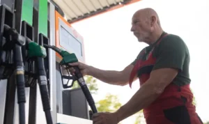 A imagem mostra um trabalhador sênior parado no posto de gasolina e no carro abastecendo. Ele usa uma blusa preta e um macacão vermelho por cima.