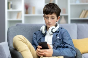 A imagem mostra um adolescente sério, com fones de ouvido sentado em casa no sofá, usando o telefone.