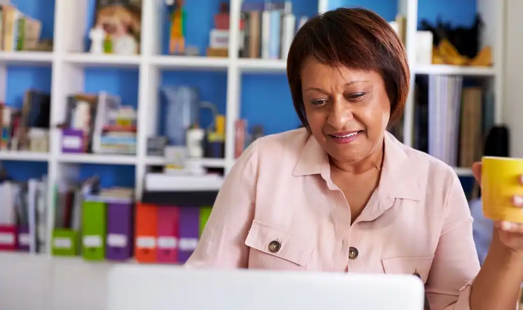 A imagem mostra uma mulher madura sorridente com laptop trabalhando no escritório doméstico. Ela usa uma blusa rosa, em tom claro e segura uma caneca amarela de café.