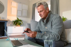 A imagem mostra um homem sênior caucasiano focado sentado no sofá, fazendo a papelada, usando smartphone e laptop.