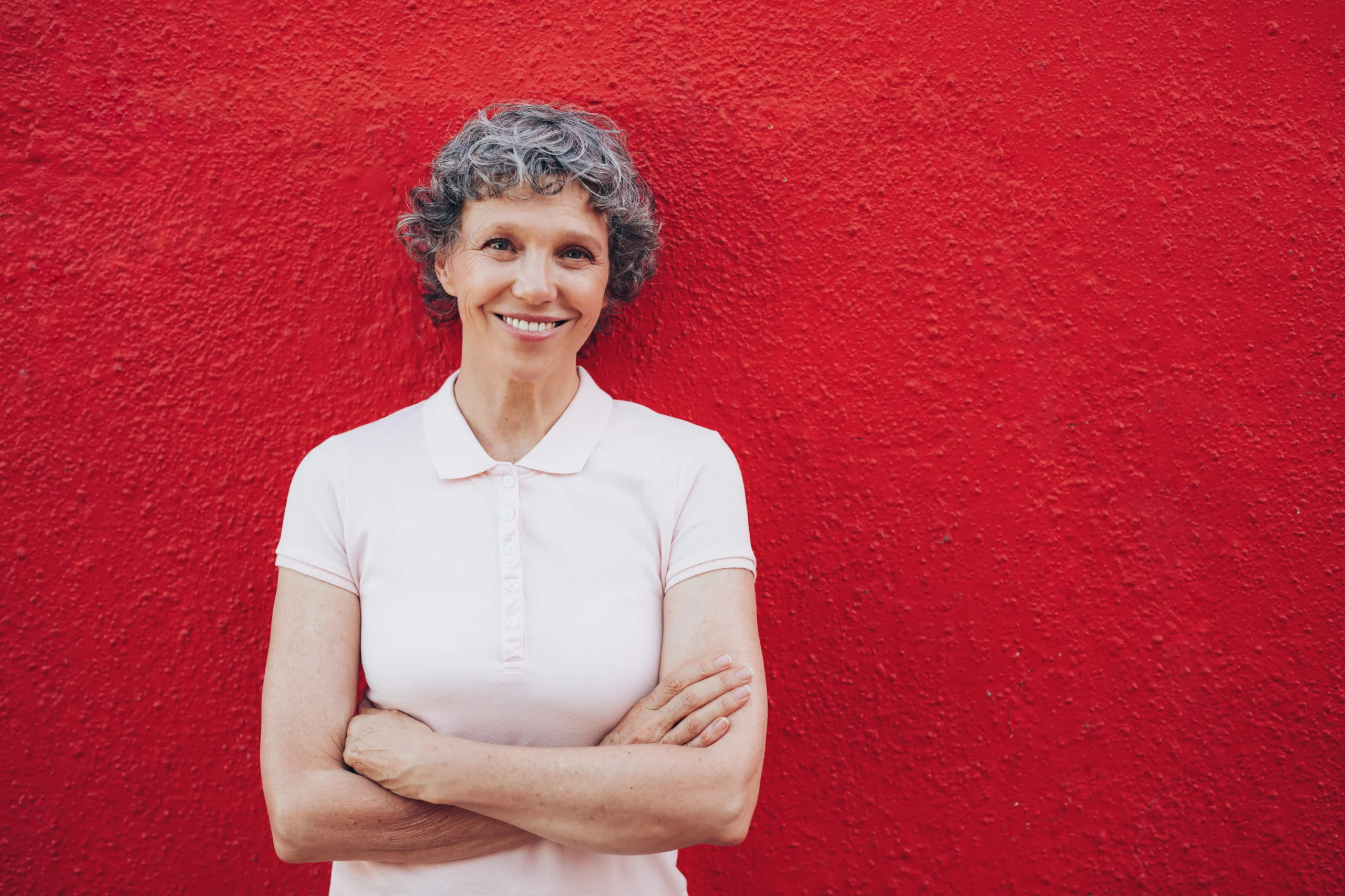 A imagem mostra uma mulher sênior, sorrindo, olhando para a câmera. Ela possui cabelos curtos grisalhos, está de braços cruzados e usa uma blusa rosa clara. Ela está em frente a uma parede vermelha.