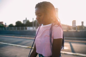 A imagem mostra uma mulher jovem, negra sozinha, luz de fundo ao ar livre, pensativa.