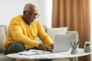 A imagem mostra um homem negro sênior digitando em laptop trabalhando on-line em um abiente fechado. Ele usa um suéter de mangas longas, amarelo e óculos de grau com aros pretos.