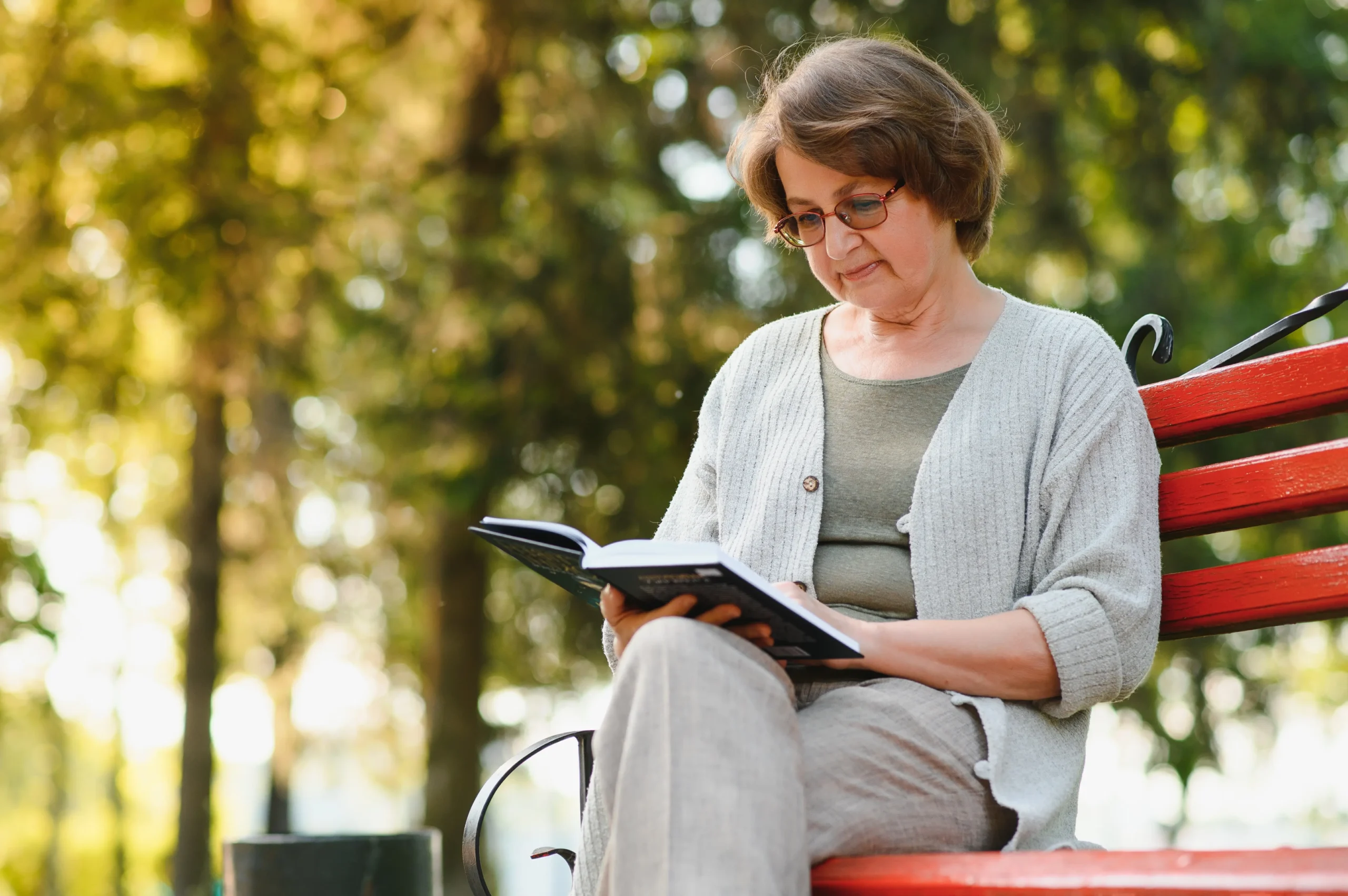 A imagem mostra uma mulher sênior feliz no parque de verão, lendo um livro e sorrindo.