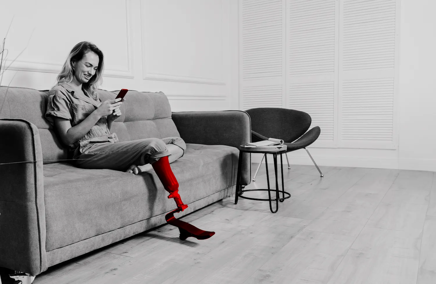 A imagem mostra uma mulher, sentada no sofá enquanto sorri e utiliza o celular. Ela possui uma prótese em uma das pernas.