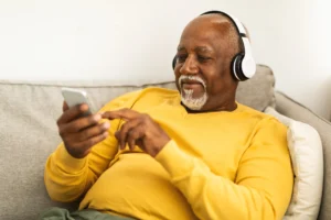 A imagem mostra um homem de meia-idade, sorrindo, sentado em um sofá, usando fones de ouvidos e usando um smartphone.