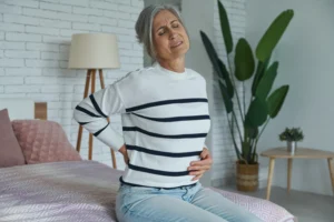 A imagem mostra uma mulher sênior, sofrendo de dor nas costas, enquanto está sentada na cama em casa. Ilustra o texto sobre hérnia de disco aposenta.