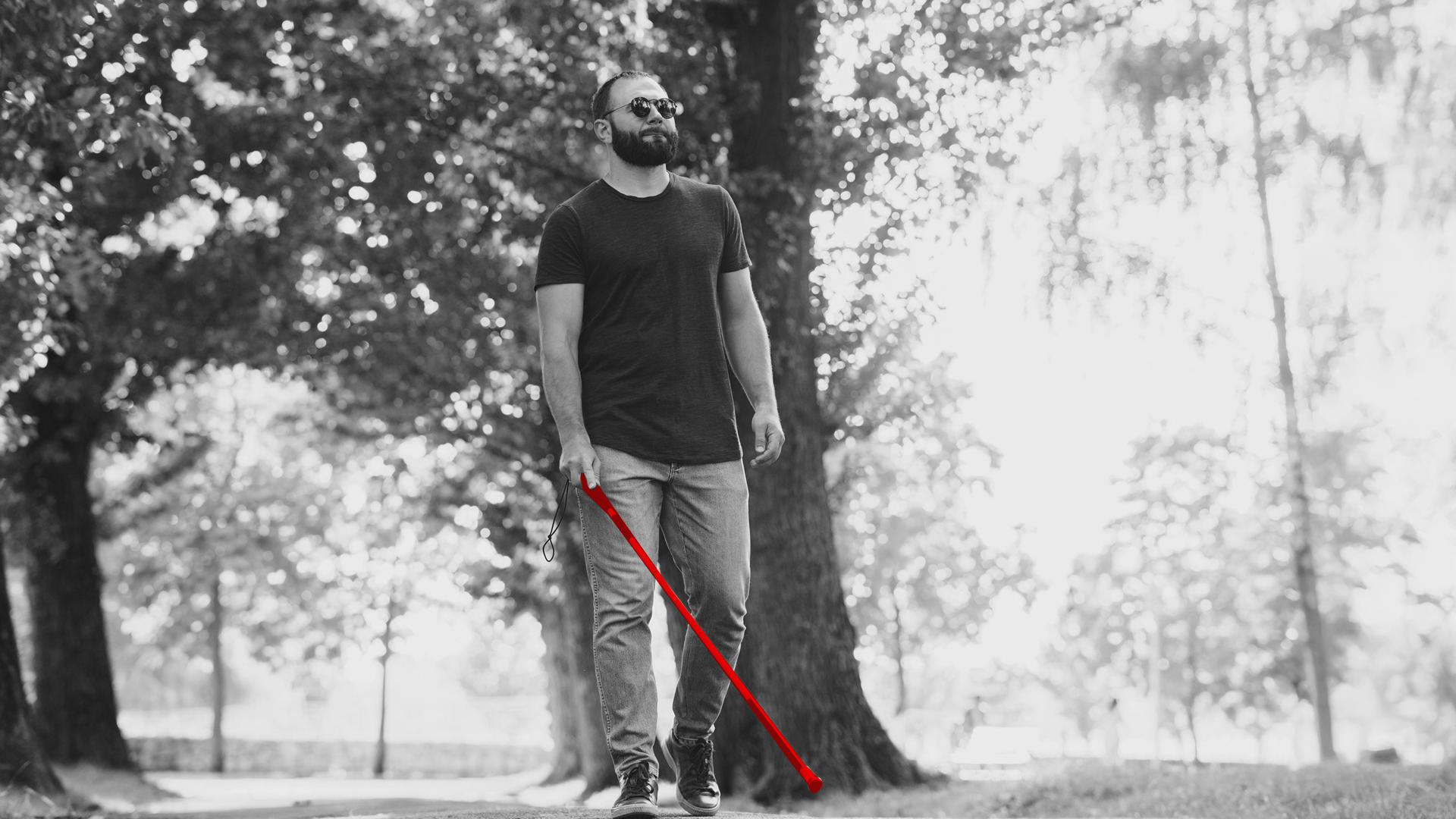 A imagem mostra um homem cego, com bengala e óculos, passeando em ar livre.