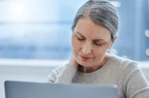 A imagem mostra uma mulher sênior, usando o laptop e com dores no pescoço. Ilustra o texto sobre osteoporose.