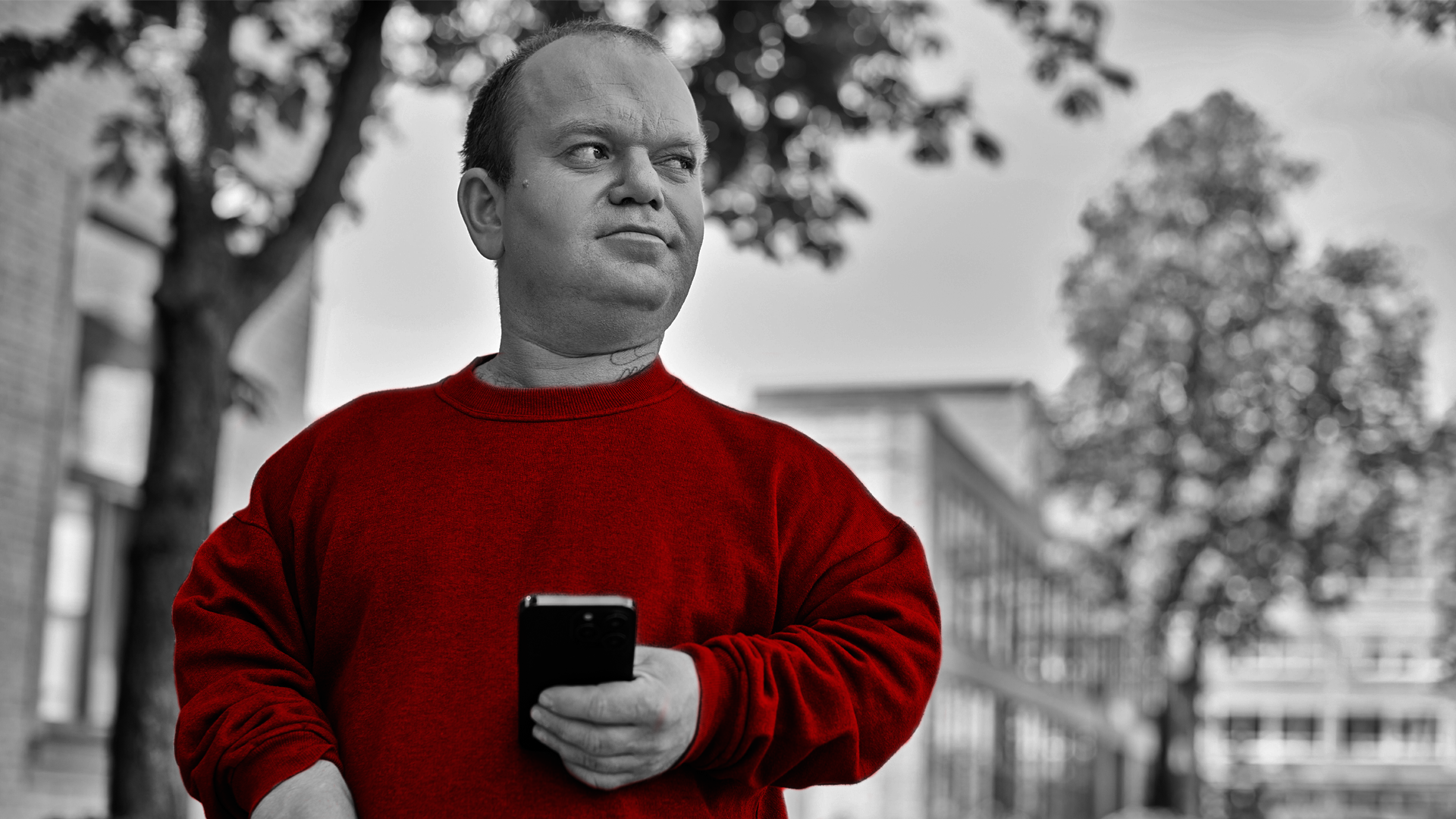 A imagem mostra um homem PcD, com nanismo, segurando com uma das mãos um smartphone, ao ar livre.