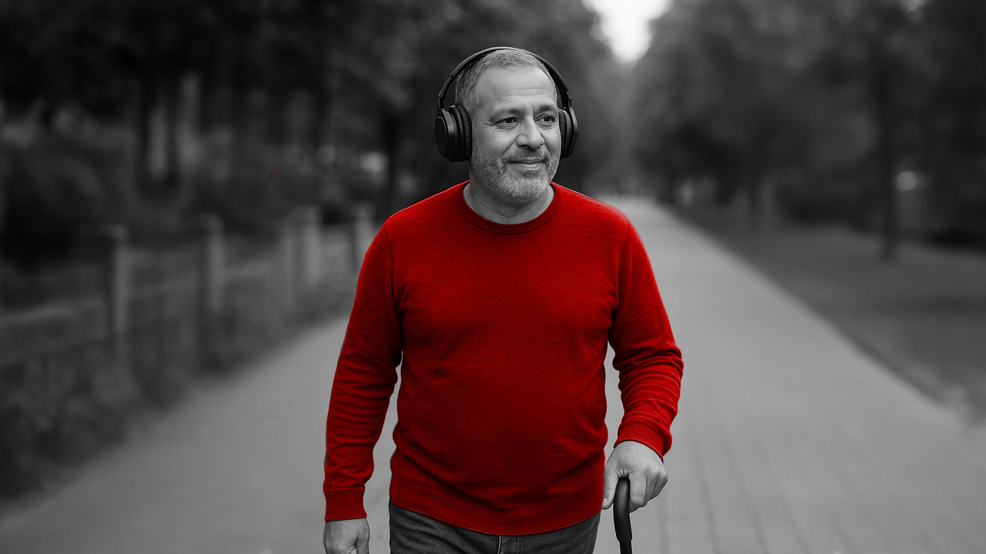 A imagem mostra um homem de meia-idade, sorrindo, andando ao ar livre, com uma bengala e fones de ouvido (abafadores).