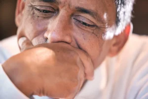 A imagem mostra em destaque, o rosto de um homem de meia-idade, reflexivo, com uma das mãos na boca.