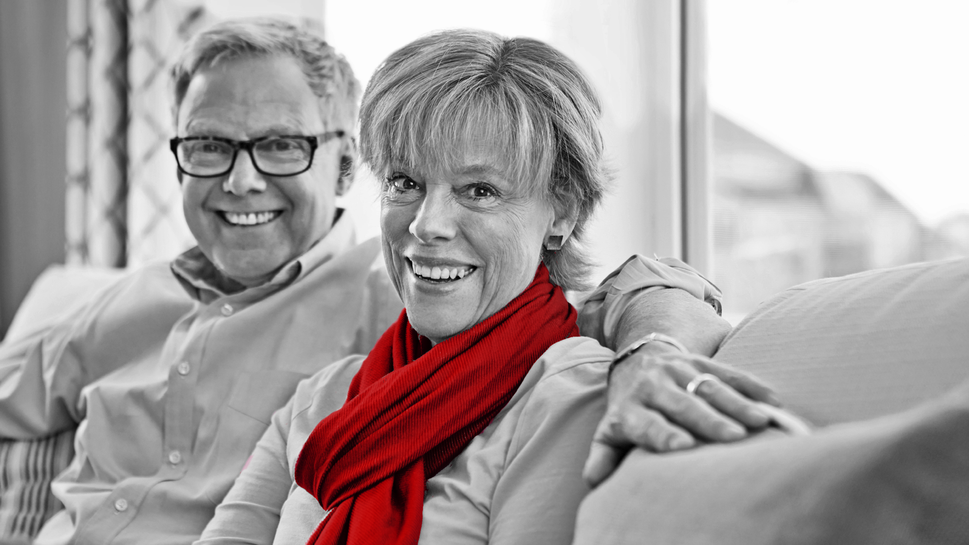 A imagem mostra um casal maduro, sentando em seu sofá, dentro de casa. Ambos sorriem para a câmera.