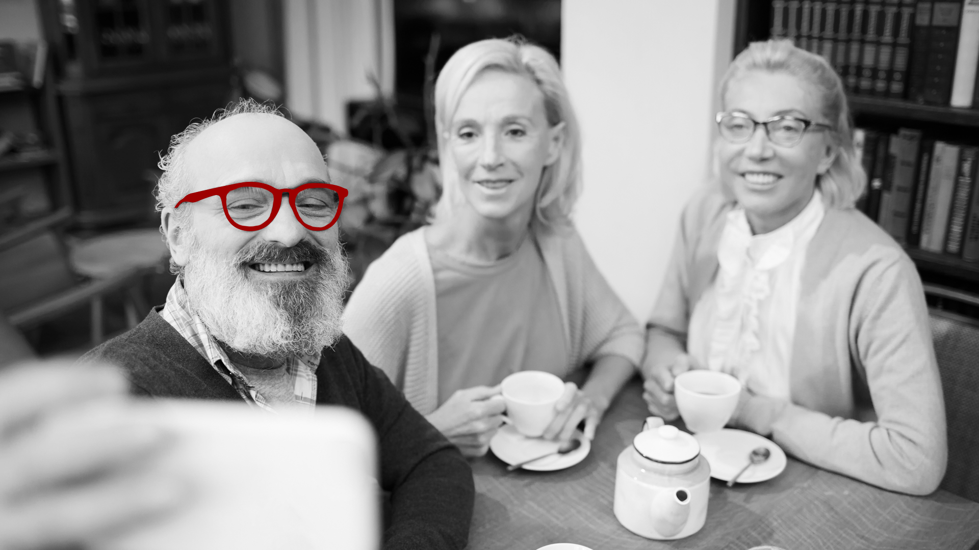 A imagem mostra 3 amigos maduros, em um café, sentados à mesa. O homem está apontando o celular, para tirar uma selfie e todos estão sorrindo.