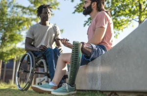 A imagem mostra dois amigos, conversando ao ar livre. Um deles é cadeirante e, o outro, tem uma prótese em uma das pernas. Ilustra o texto sobre aposentadoria por deficiência física.