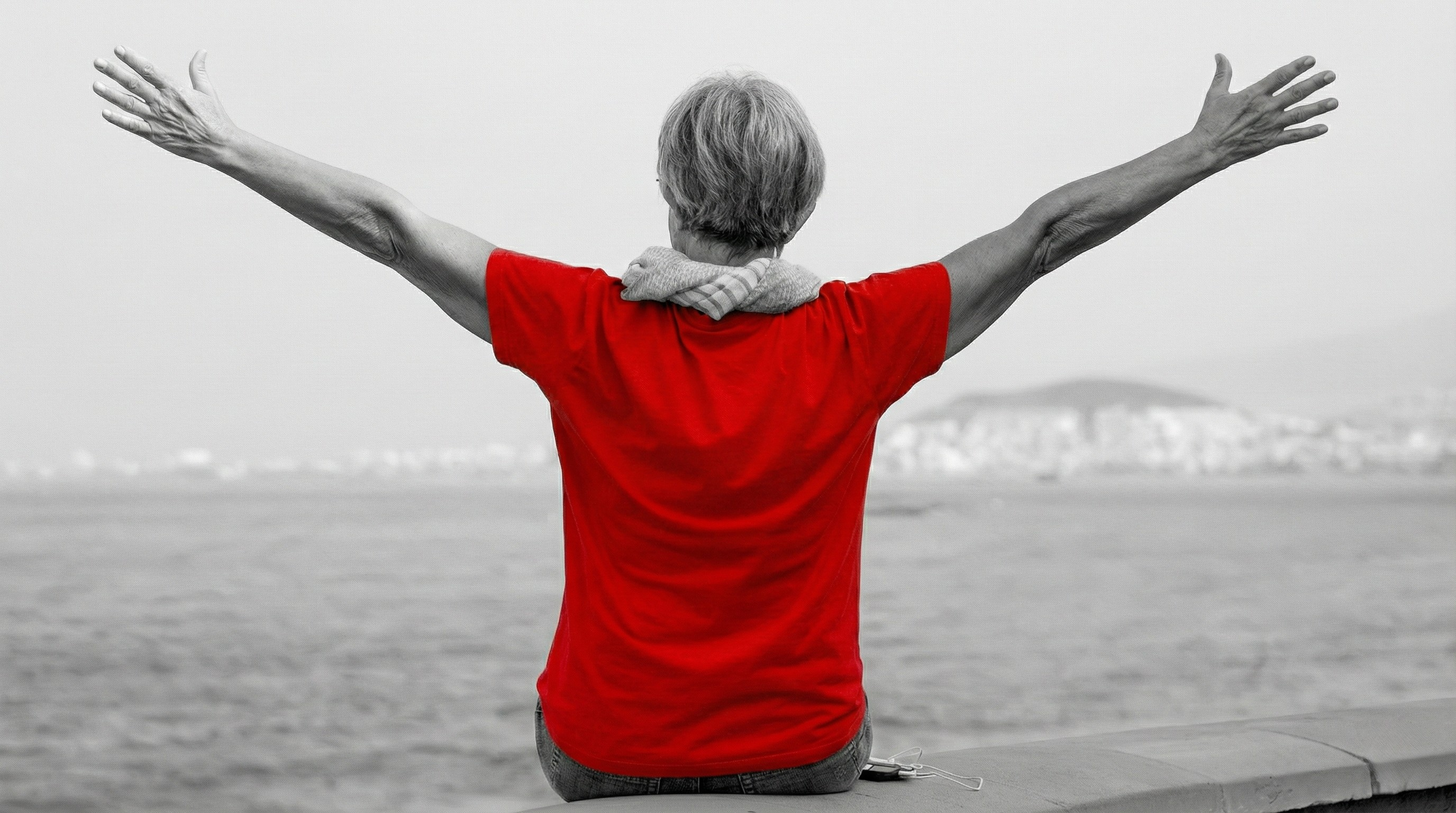 A imagem mostra uma mulher de meia-idade, de costas, sentada, de braços levantados, ao ar livre.