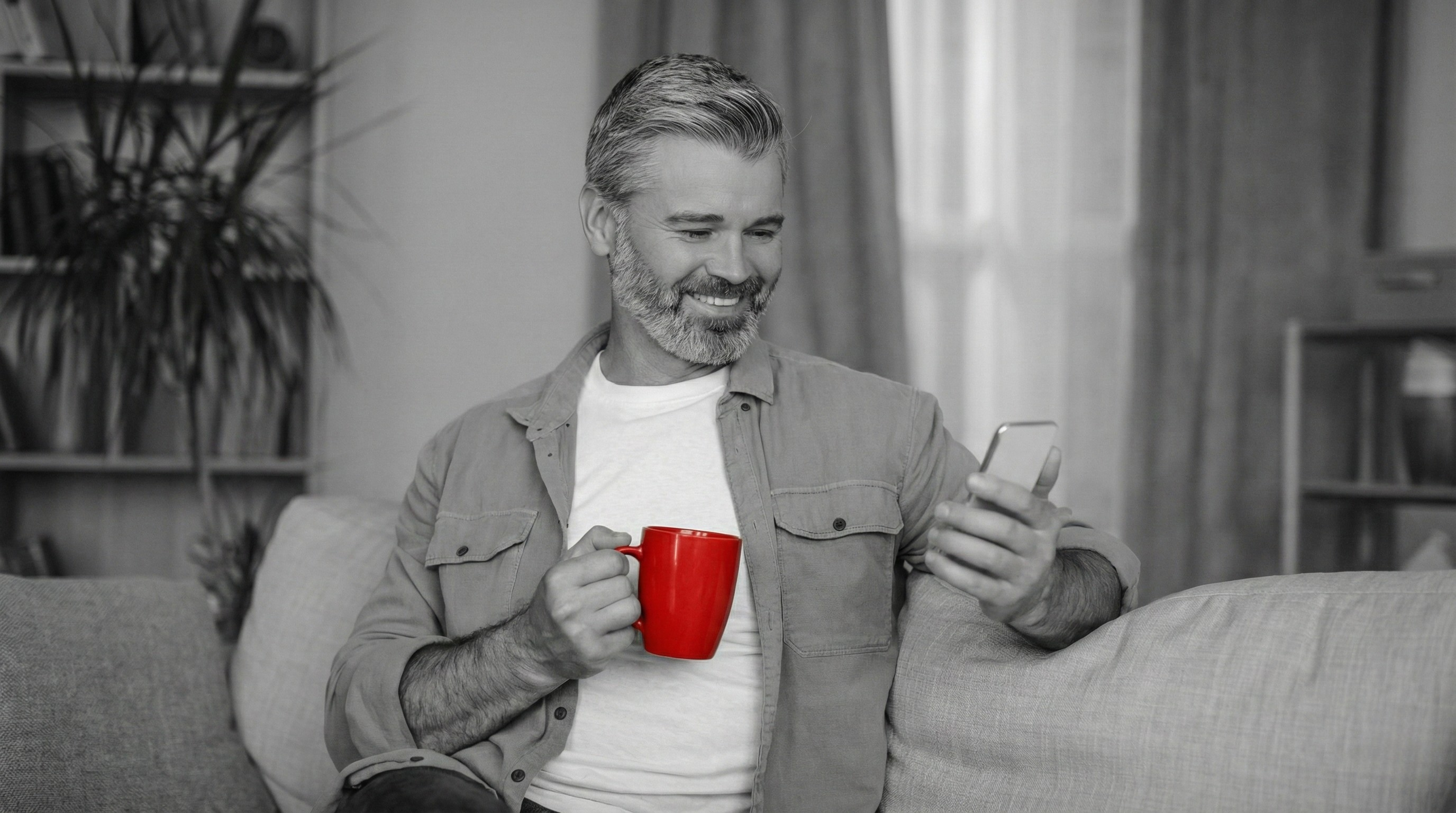 A imagem mostra um homem de meia-idade, sentado em um sofá, dentro de casa, sorrindo. Ele segura um smartphone com uma das mãos e, com a outra, uma caneca.