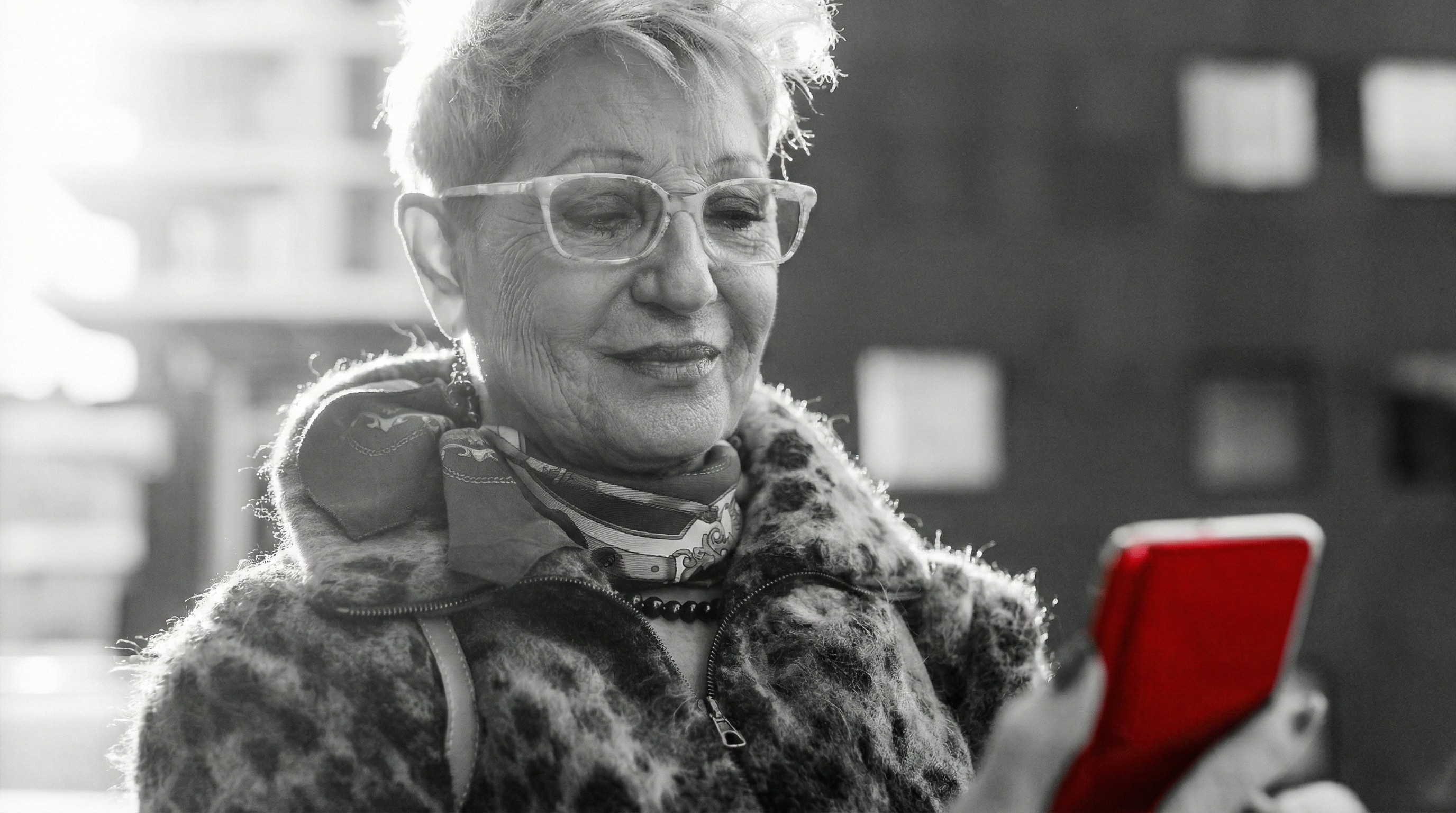 A imagem mostra uma mulher idosa, ao ar livre, sorrindo, enquanto segura um smartphone com a mão.