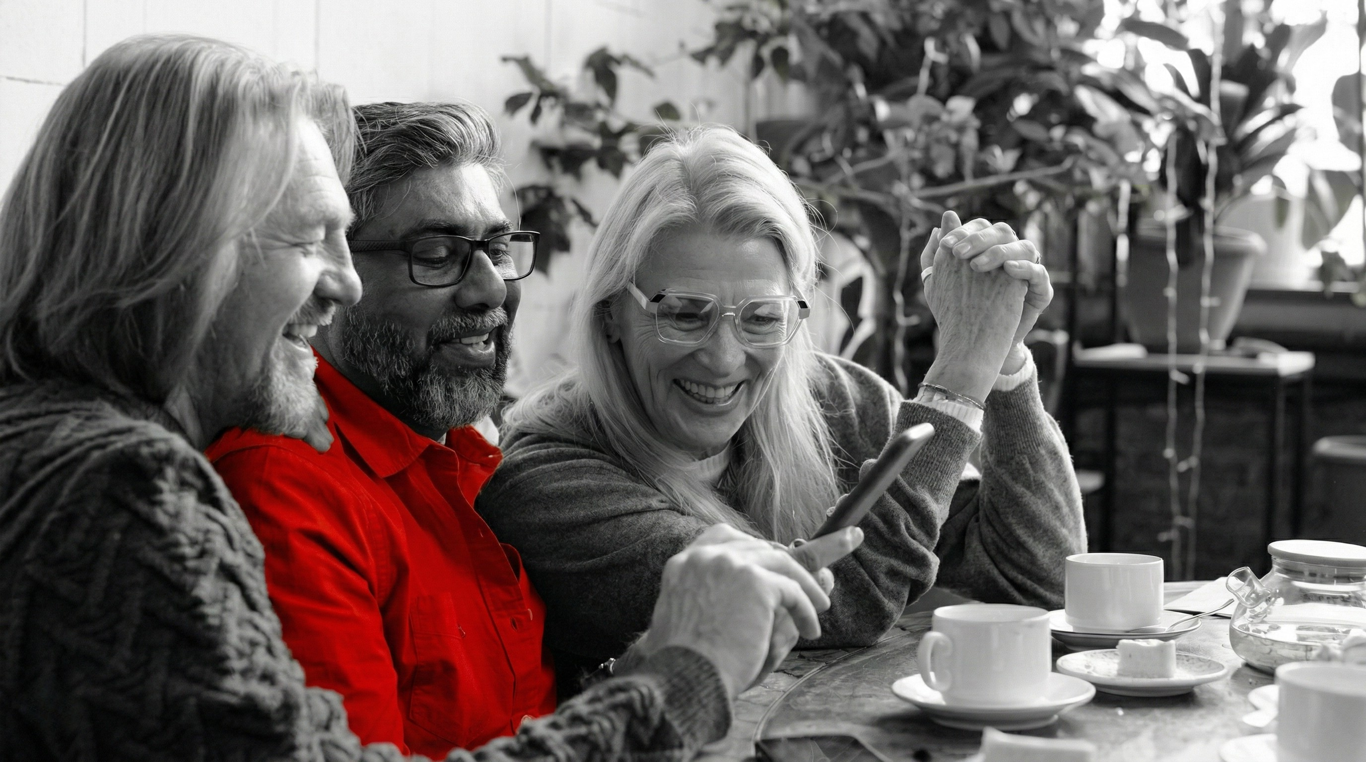 A imagem mostra um trio de amigos de meia-idade, sorrindo juntos à mesa.