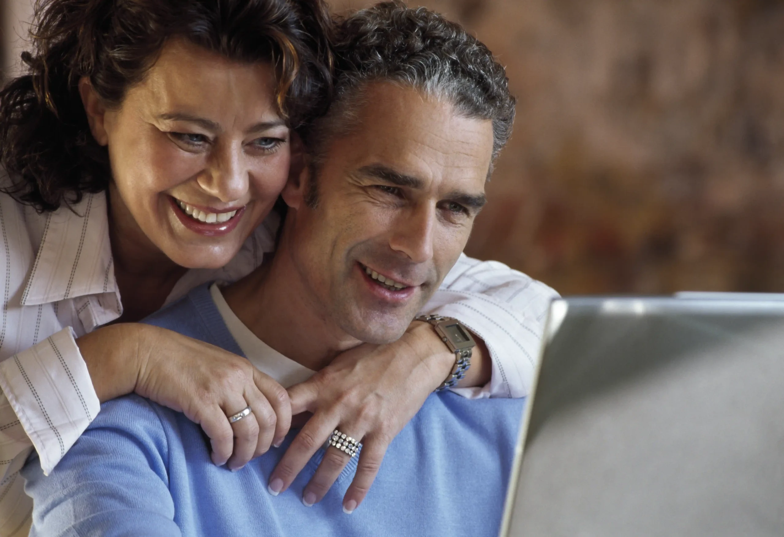 A imagem mostra um casal de meia-idade, utilizando o laptop juntos, enquanto sorriem. Ilustra o texto sobre tabela da aposentadoria.