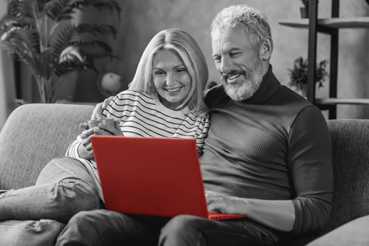 A imagem mostra um casal de meia-idade, sentado no sofá de casa, enquanto olham para o laptop que está no colo do homem.
