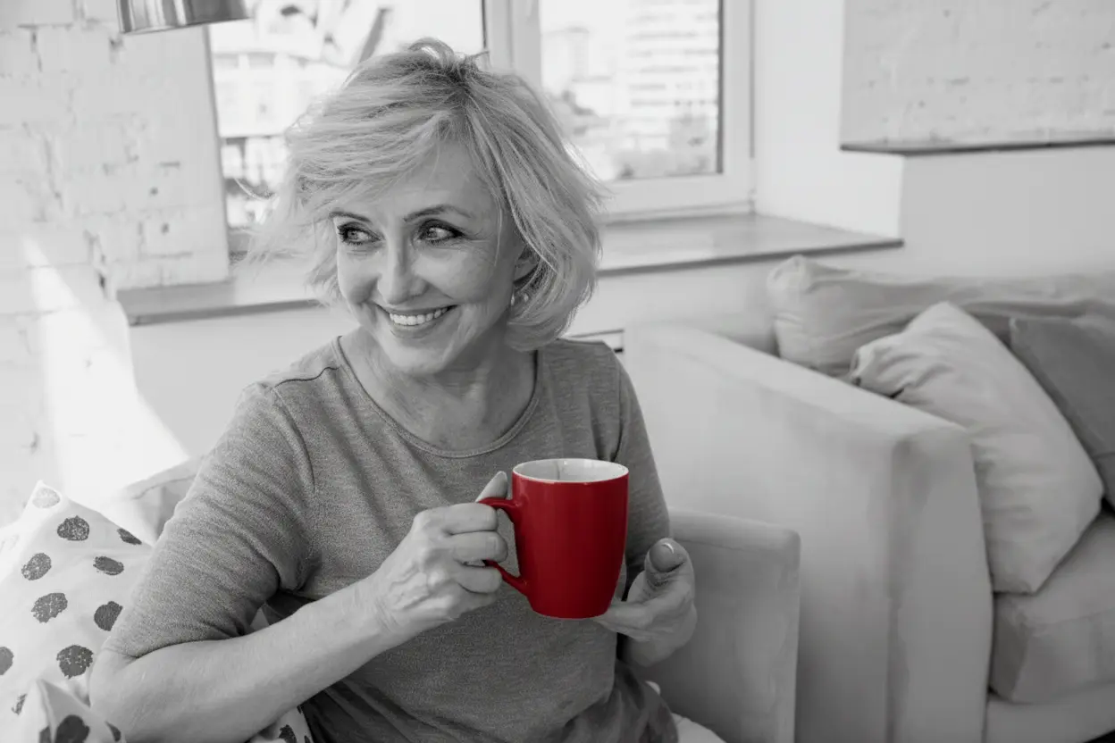 A imagem mostra uma mulher de meia-idade, sentada no sofá de casa, sorrindo, enquanto segura uma xícara.