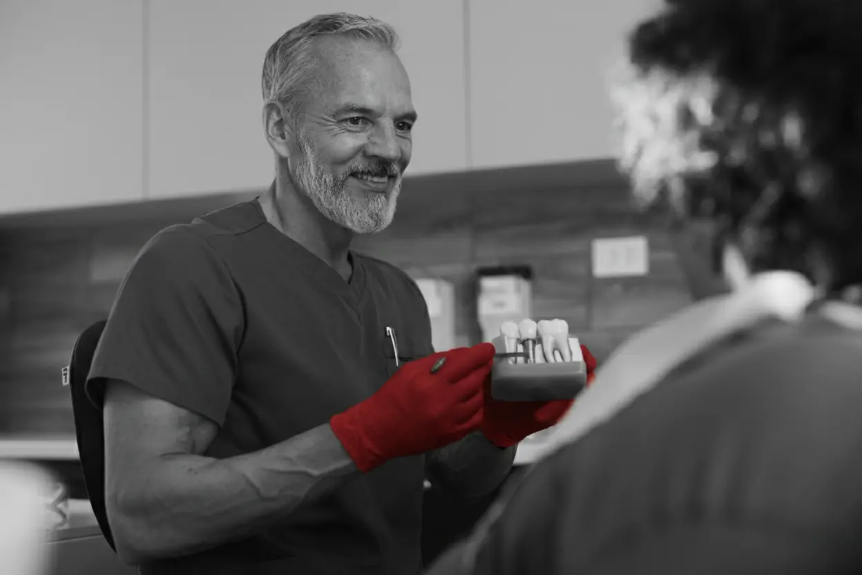 A imagem mostra um dentista de meia-idade, de roupas cirúrgicas e luvas, atendendo uma paciente, enquanto sorri.