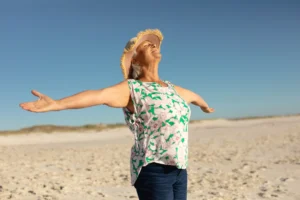 A imagem mostra uma mulher idosa, de abraços abertos, curtindo estar em uma praia, ao ar livre. Ilustra o texto sobre IRPF para aposentados .