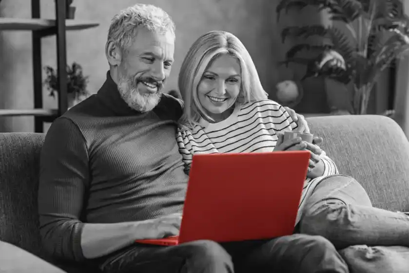 A imagem mostra um casal de meia-idade, sentado no sofá de casa, enquanto olham para o laptop que está no colo do homem.