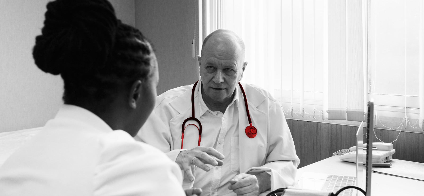 A imagem mostra um médico sênior, usando jaleco e estetoscópio, em seu consultório, consultando uma paciente.