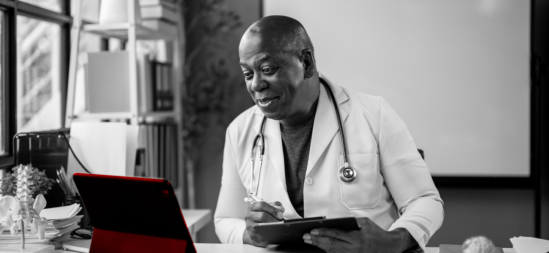 A imagem mostra um médico de meia-idade, usando jaleco e estetoscópio em volta do pescoço, em seu consultório, sendo à mesa.